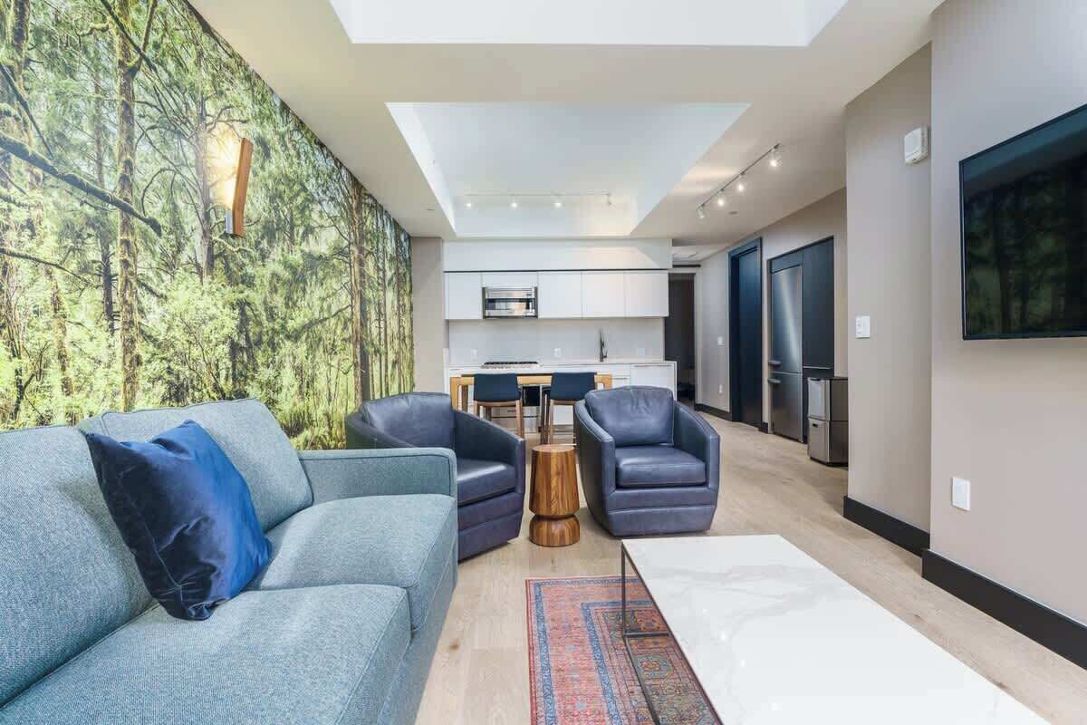 Uma sala de estar moderna com um sofá, poltronas e uma mesa de centro. É visível uma área de cozinha com armários e eletrodomésticos ao fundo.