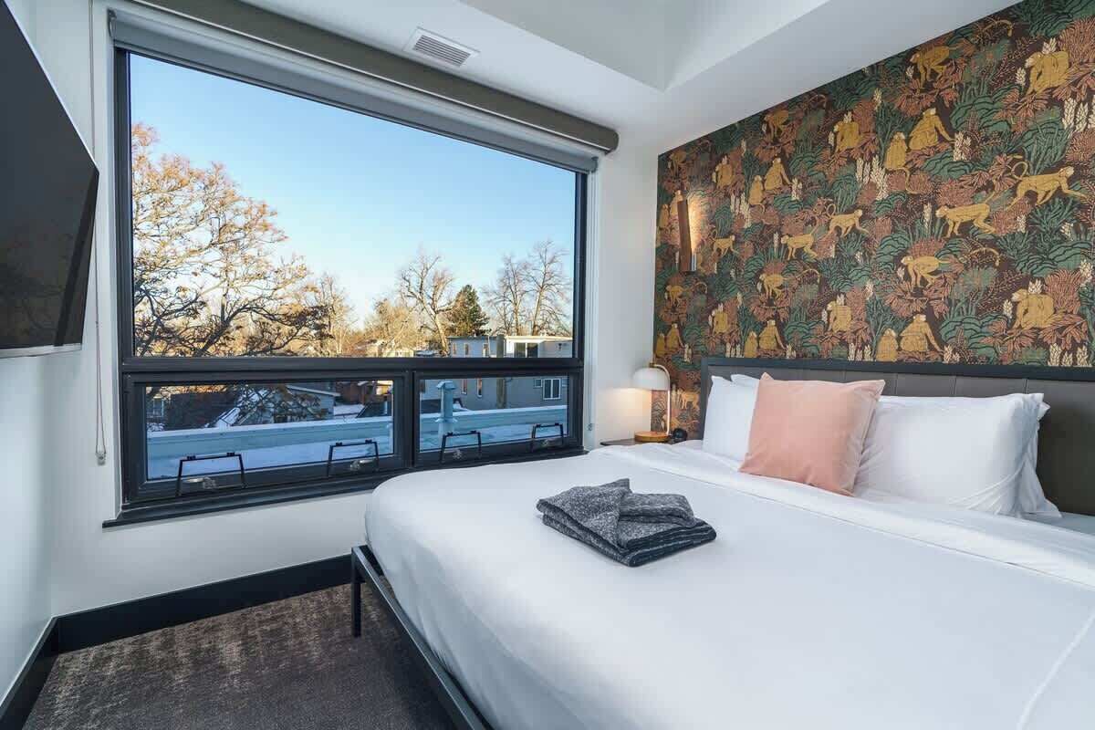 Une chambre d’hôtel avec une grande fenêtre, un lit agrémenté d’oreillers blancs et roses, et un mur orné d’une tapisserie aux motifs élaborés.