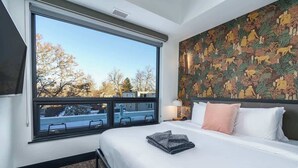 Une chambre d’hôtel avec une grande fenêtre, un lit agrémenté d’oreillers blancs et roses, et un mur orné d’une tapisserie aux motifs élaborés.