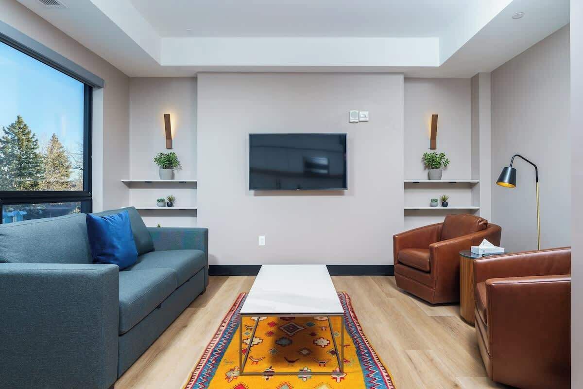 Una sala de estar moderna con un sofá gris, un sillón de cuero marrón y una mesa de centro blanca. Hay un televisor de pantalla plana montado en la pared, y una mesita de noche con una lámpara a la derecha.