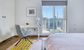 A hotel room with a bed, a desk, a chair, a window with a city view, and a framed picture on the wall.