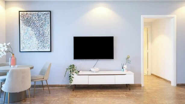 A modern living room with a flat-screen TV mounted on the wall, a white console table, a dining area with a round table and chairs, and a large abstract painting.