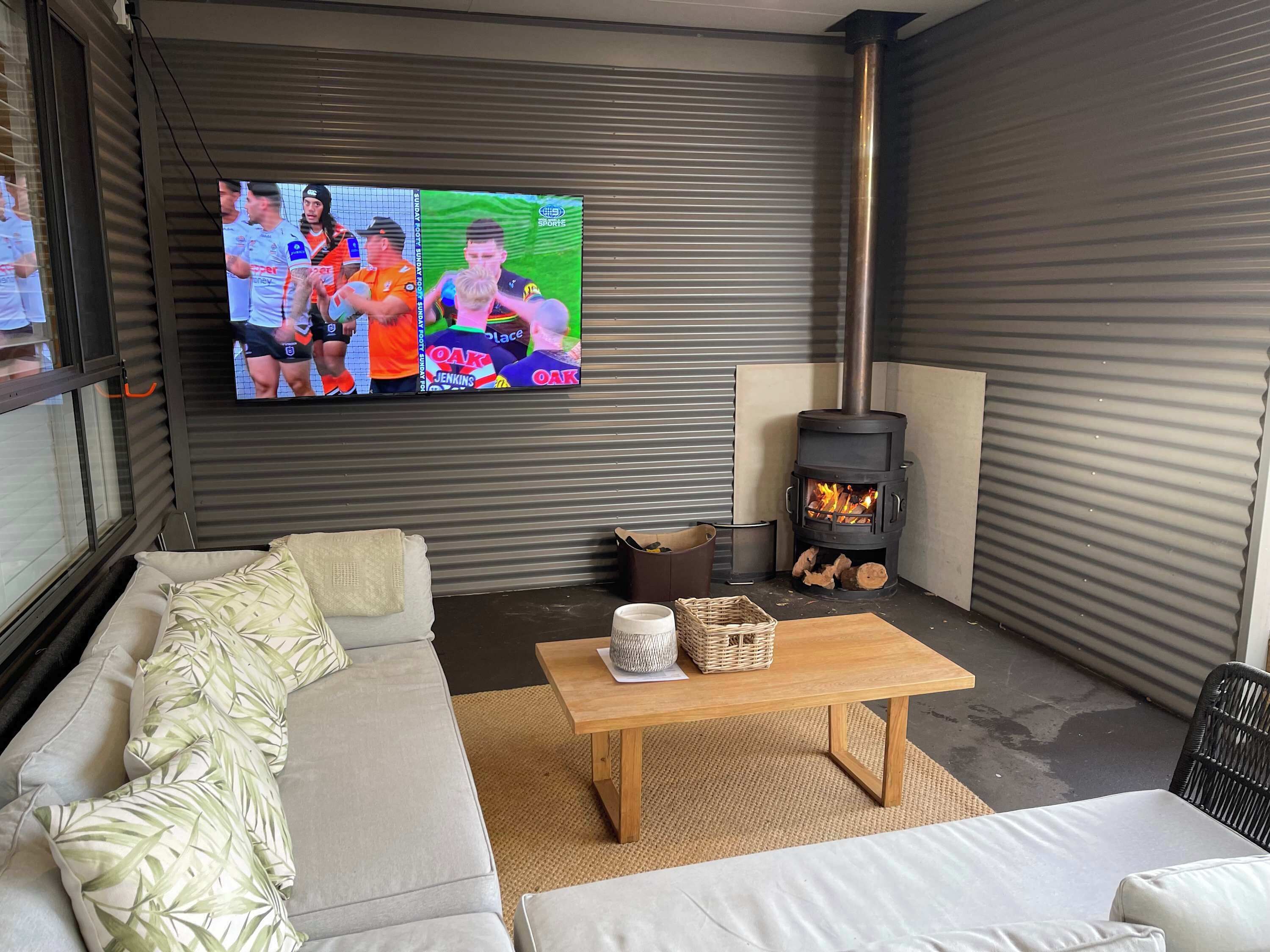 Een moderne woonkamer met een bank, een houten tafel, een tv waarop sport wordt vertoond en een houtkachel.