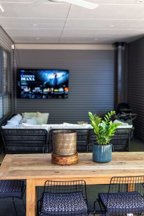 A dining area with a wooden table, black wire mesh chairs, a potted plant, a metal perforated container, and a television in the background. - Dalton Cottage - Charm & Convenience (Orange)
