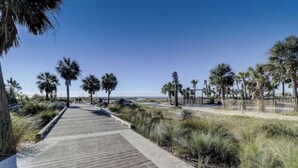 Un paseo marítimo de madera que lleva a una playa con palmeras y cielo despejado.