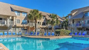 Una piscina con sillones reclinables azules y palmeras, rodeada de edificios de varios pisos con balcones.