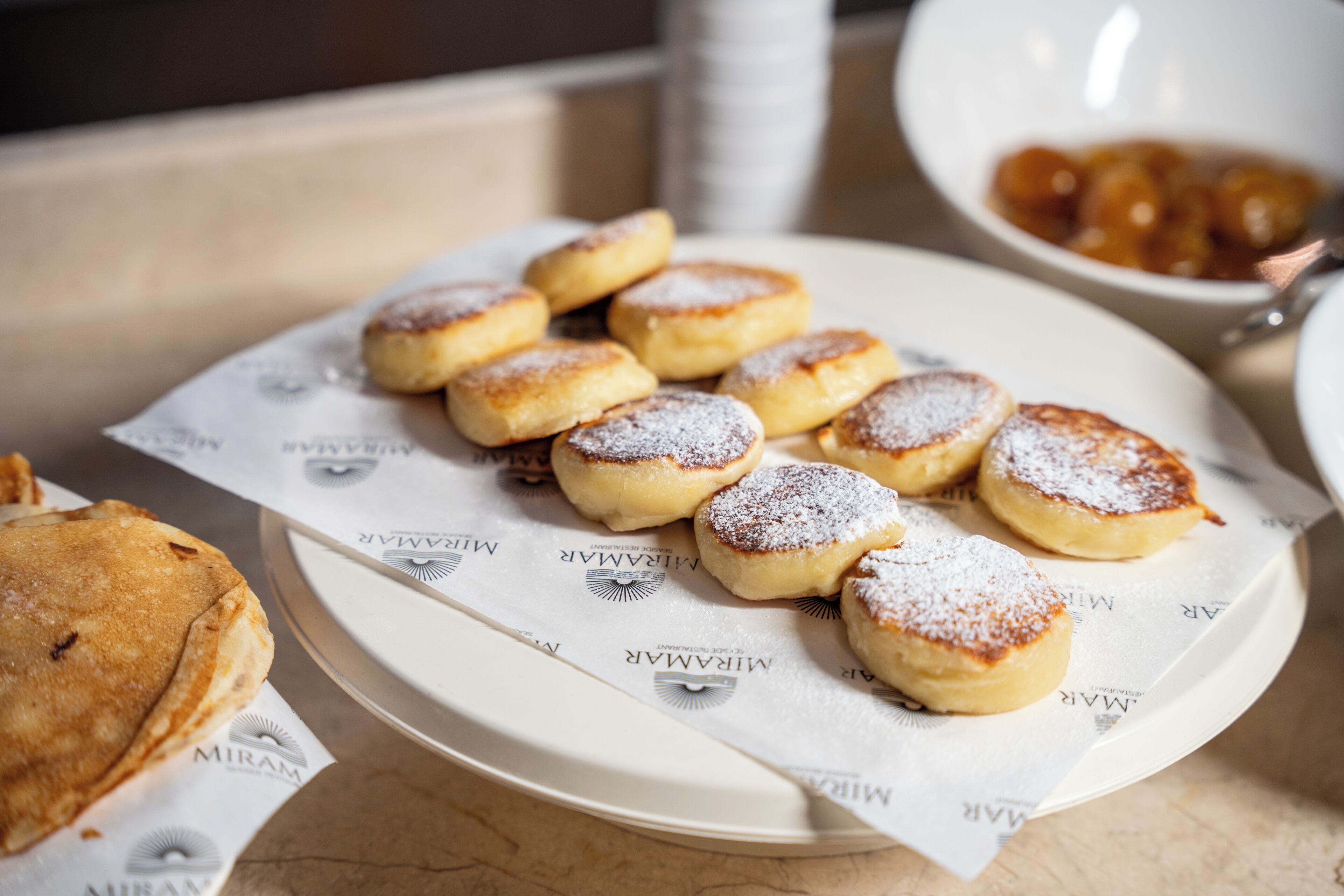 A plate of small, round pastries dusted with powdered sugar, a bowl of fruit, and a stack of white plates on a table.