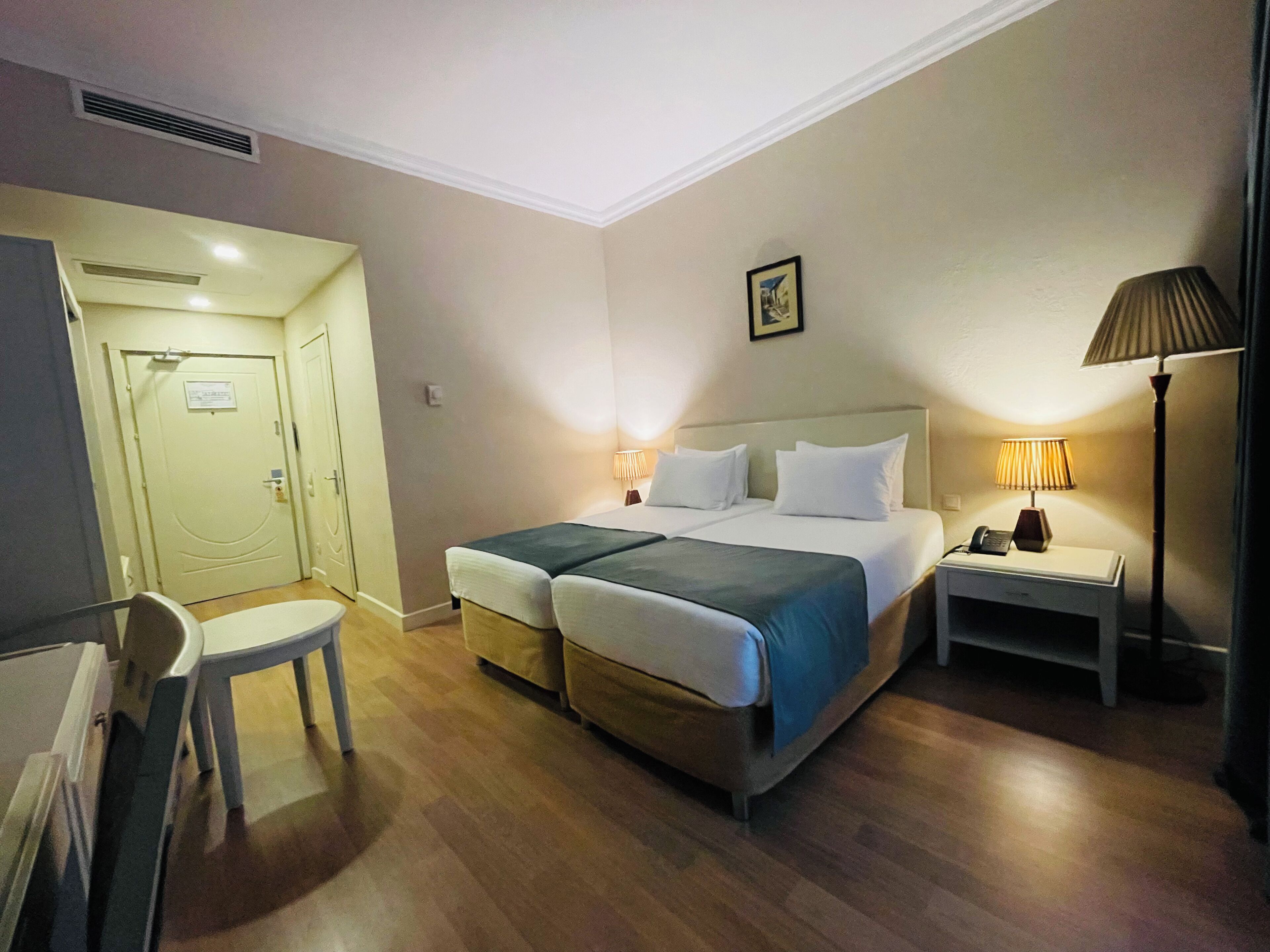 A hotel room with a double bed, a desk with a lamp, a chair, and a door.