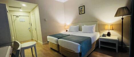 A hotel room with a double bed, a desk with a lamp, a chair, and a door.