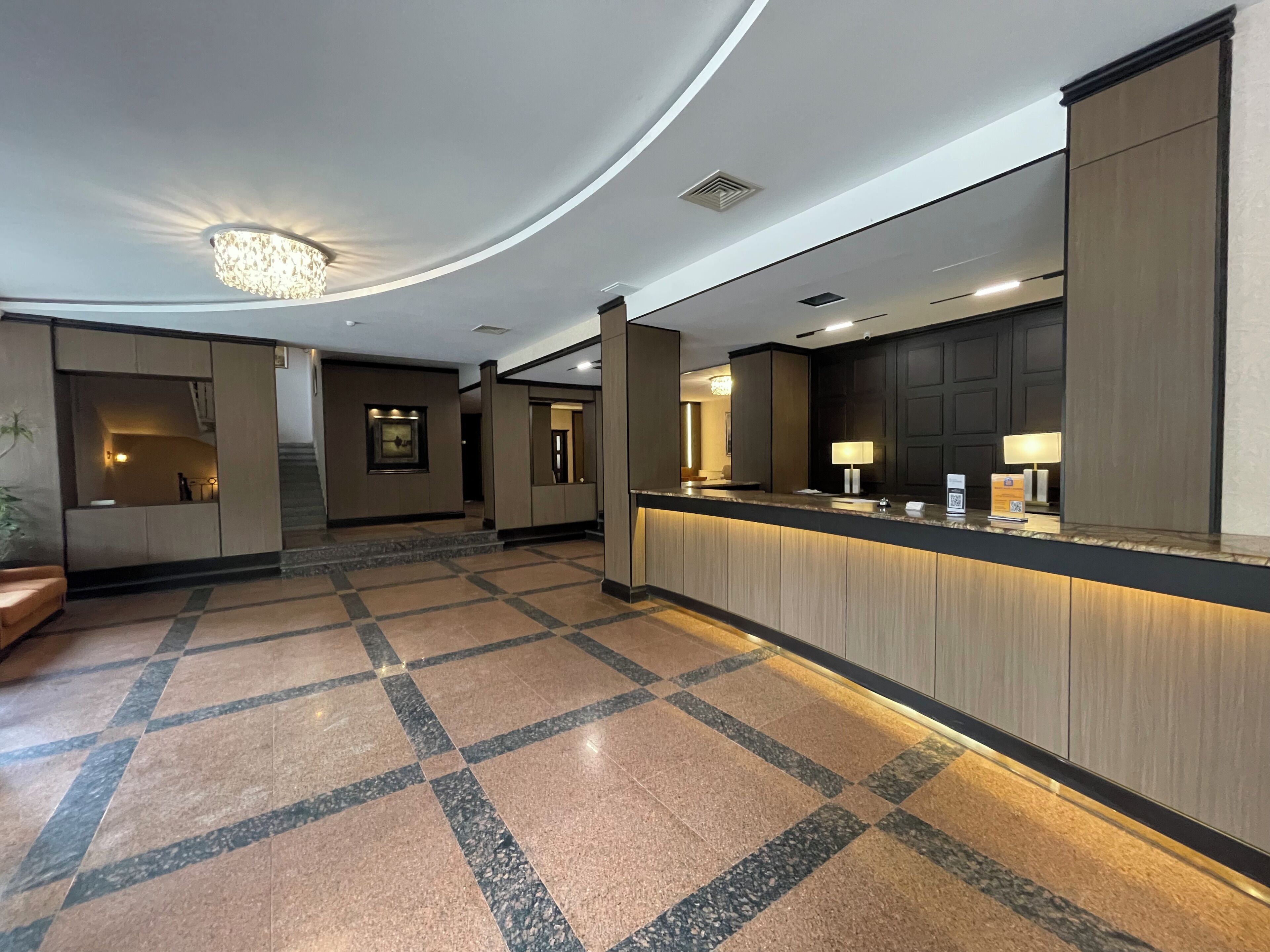 Un hall d’hôtel avec un comptoir d’accueil, un lustre, un tableau encadré au mur et un sol carrelé.