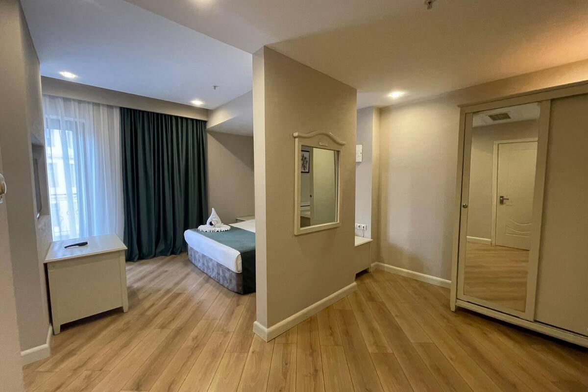 A hotel room with a bed, bedside table, mirror, wardrobe, and a window with curtains.