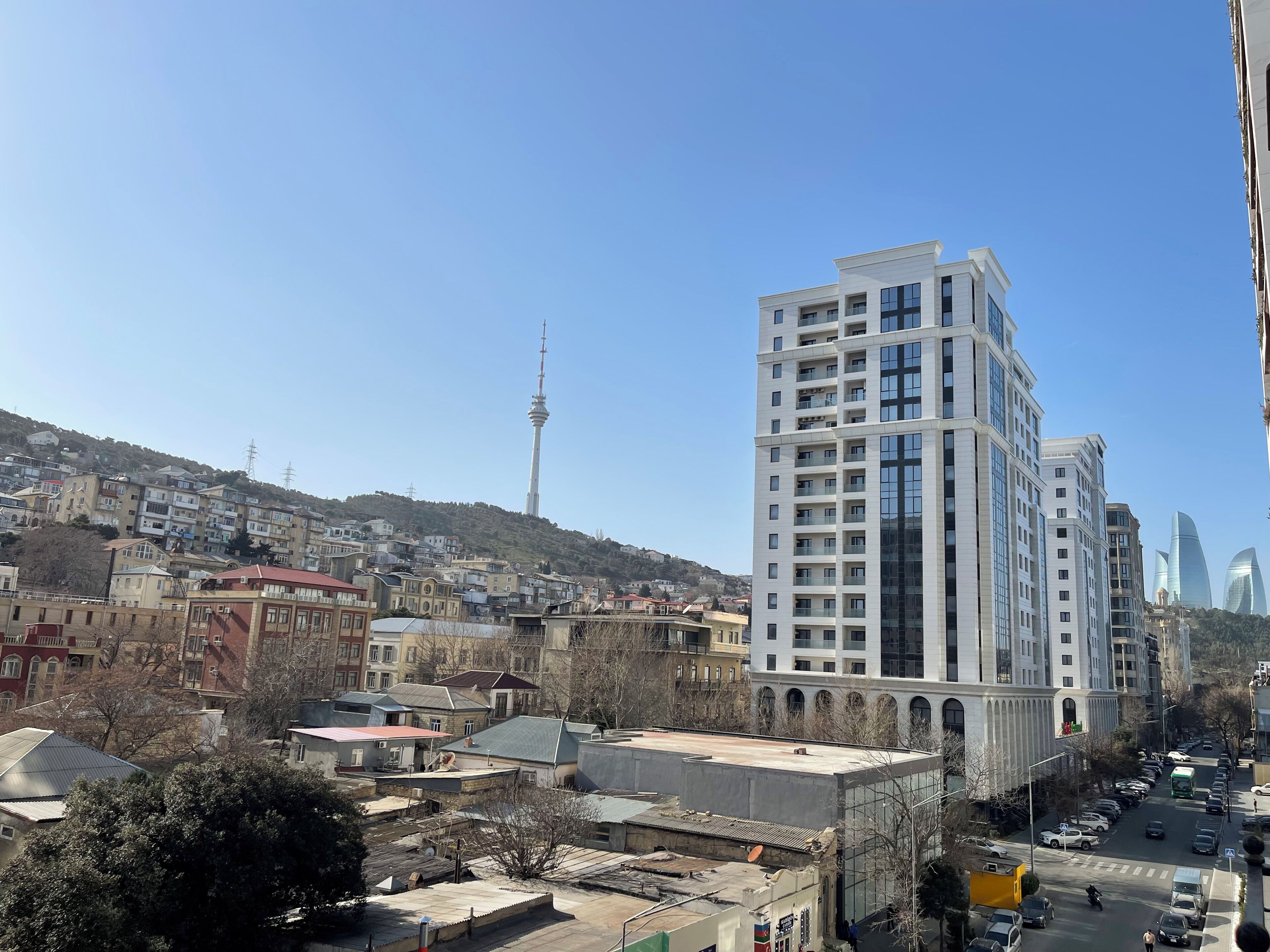 A high-rise building with a modern architectural design, a prominent tower in the background, and a street with parked cars and a few pedestrians.