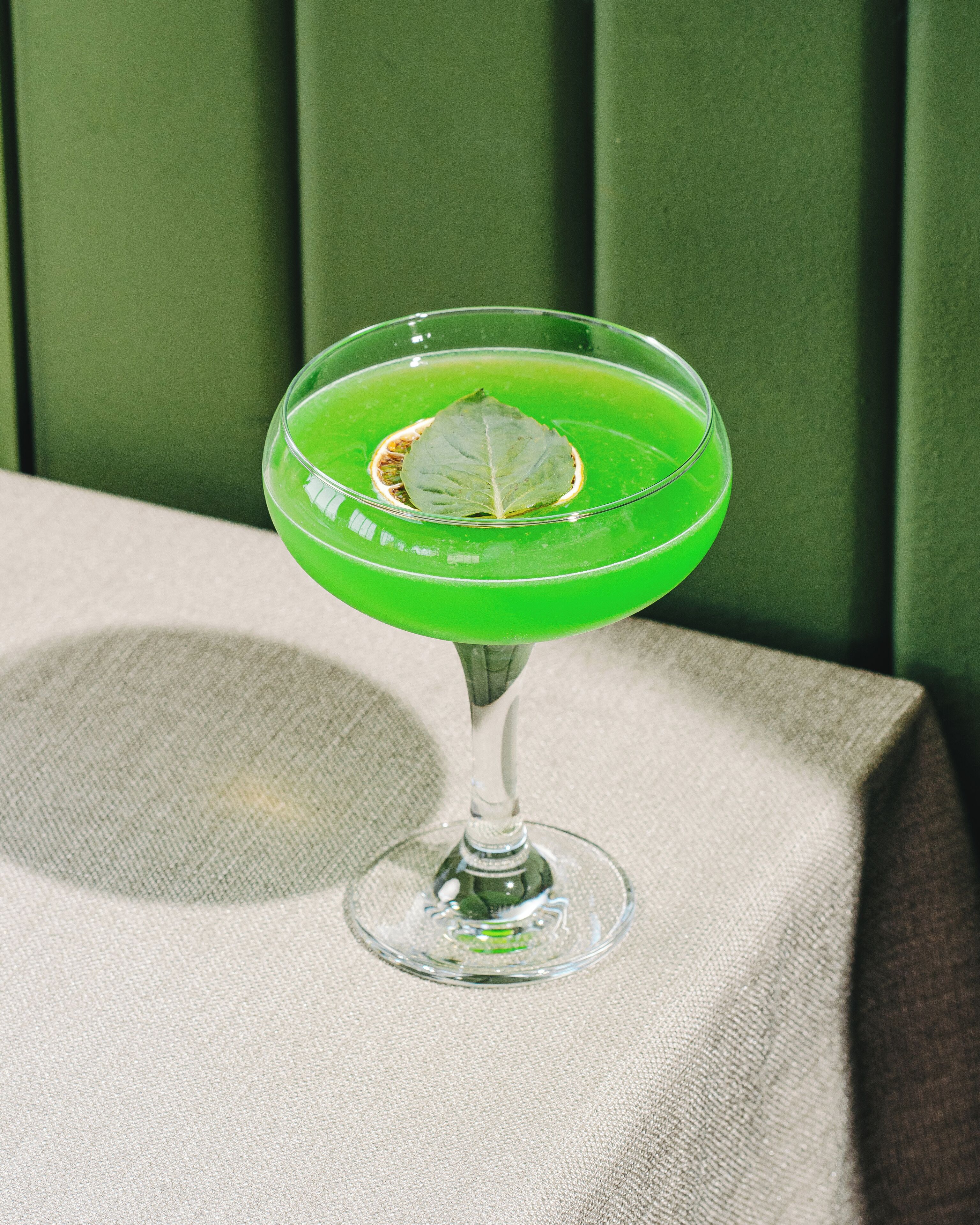 A green cocktail in a stemmed glass with a leaf garnish, placed on a textured surface.