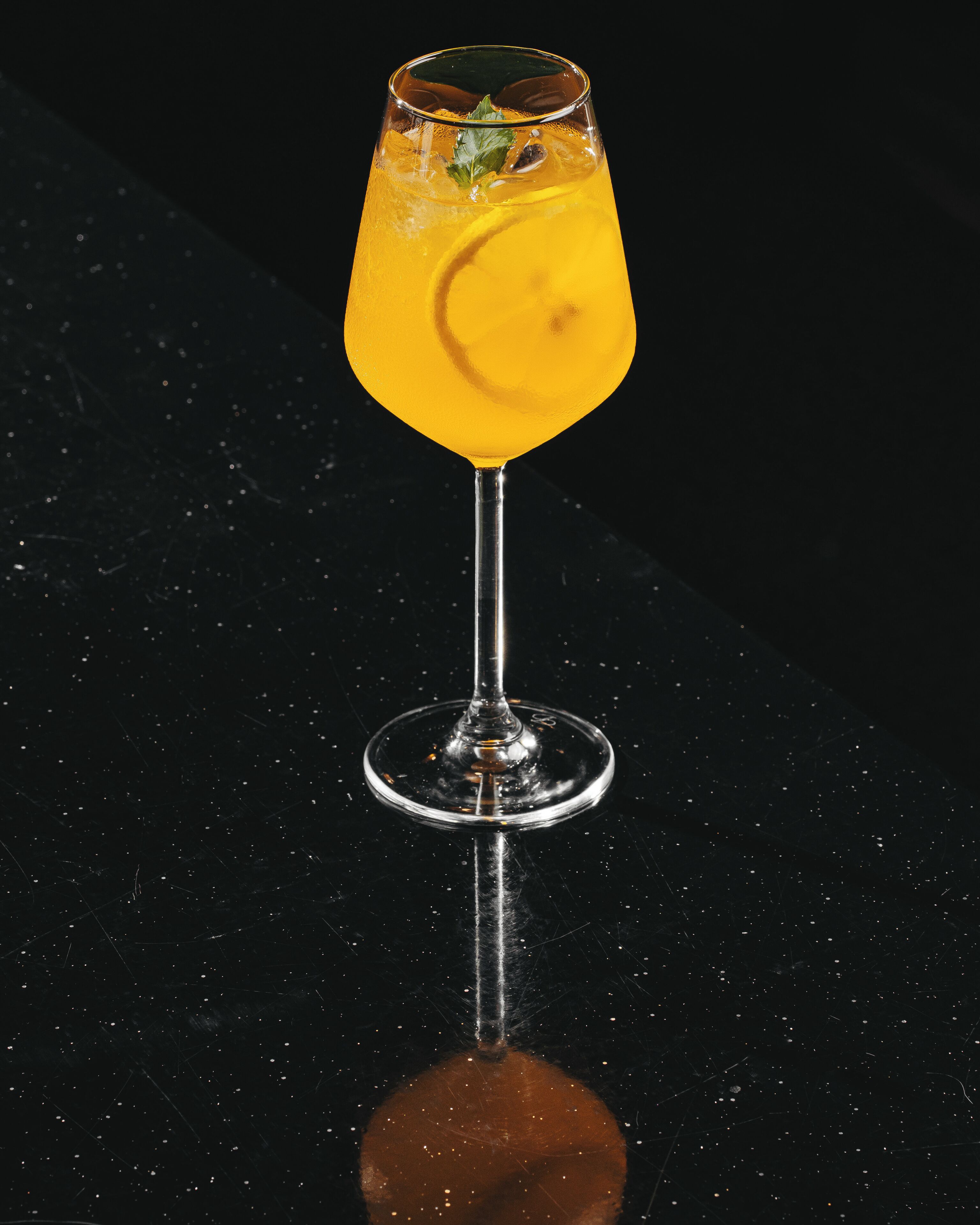 A clear glass with a yellow liquid, possibly a cocktail, with a mint garnish, set against a dark background.