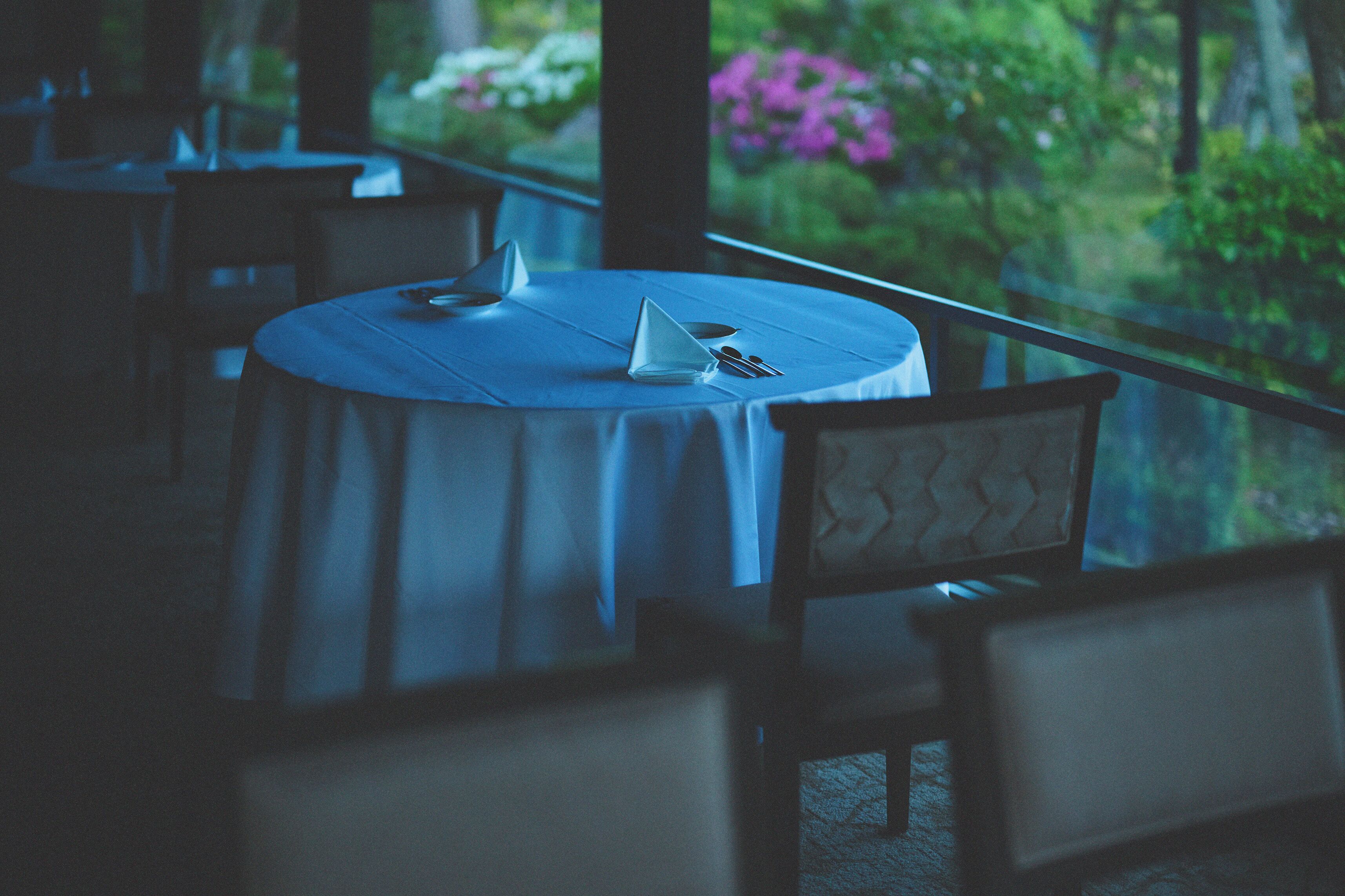 A round table with a white tablecloth, a chair with a cushioned backrest, and a view of greenery through a window.