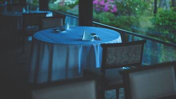 A round table with a white tablecloth, a chair with a cushioned backrest, and a view of greenery through a window.