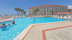 En swimmingpool med solstolar och parasoller, omgiven av palmer och en hotellbyggnad.
