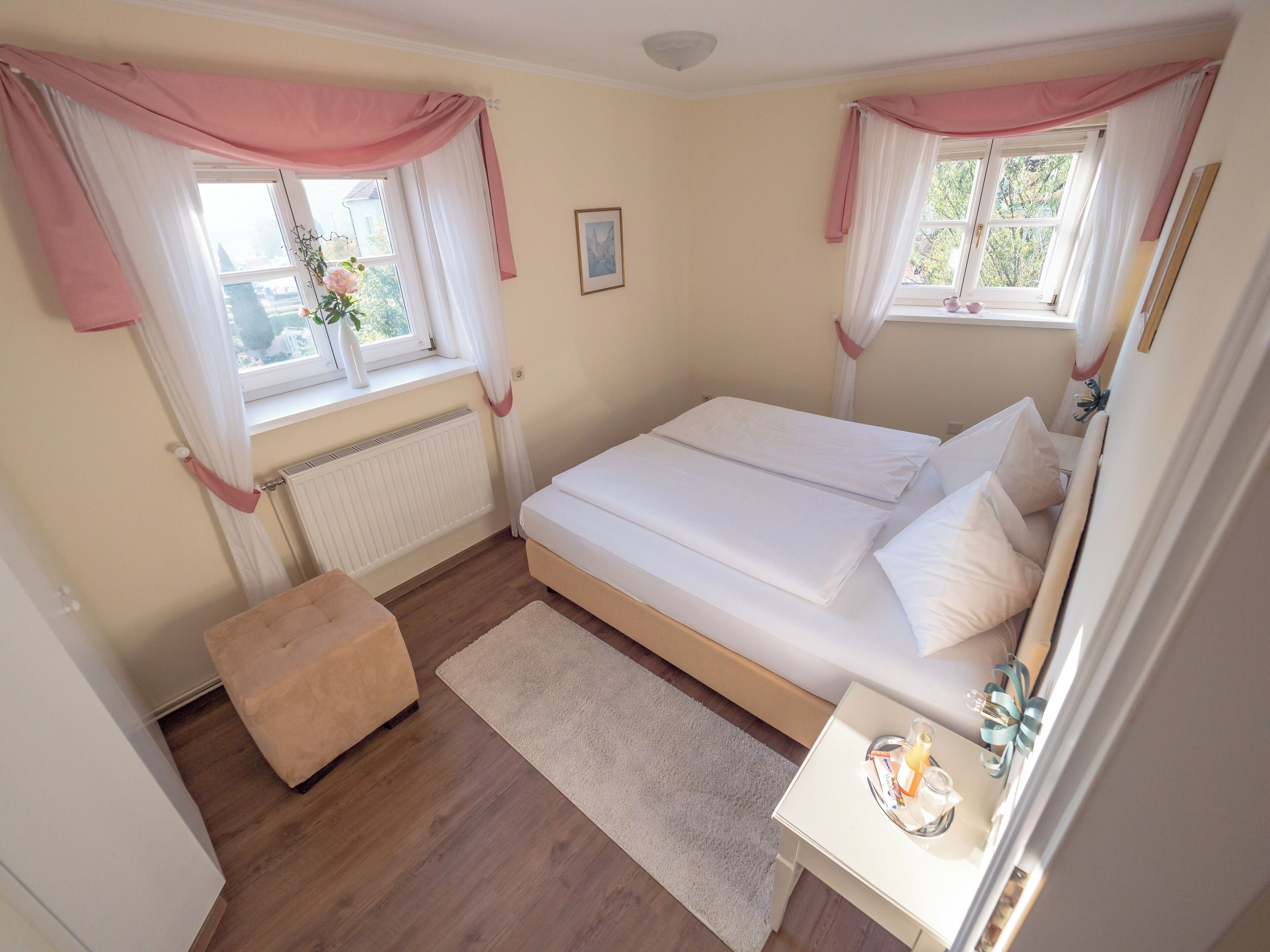 A bedroom with a bed, a bedside table, a window with curtains, a radiator, and a small stool.