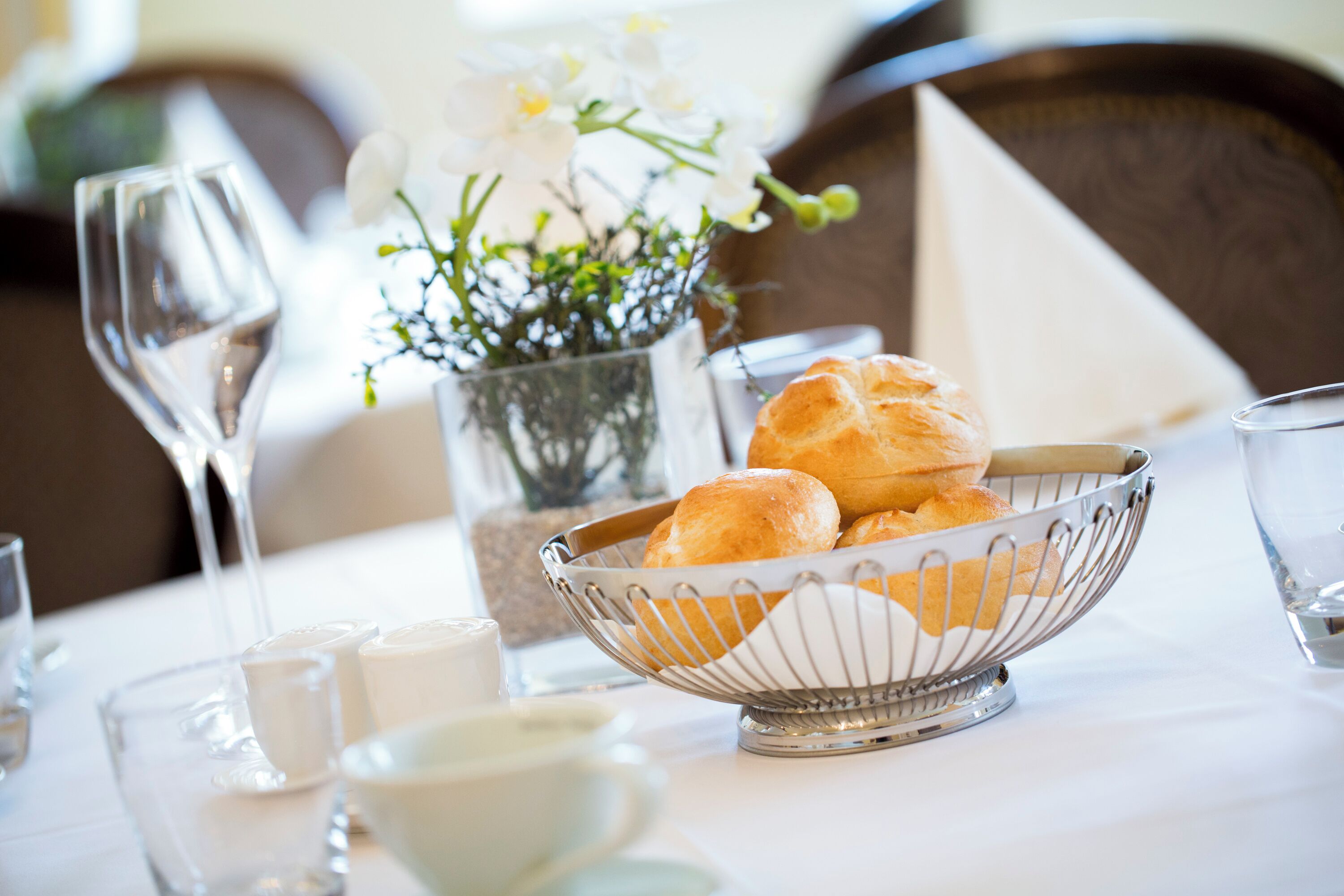 A dining table with a metal bowl containing bread rolls, a vase with white flowers, and several glasses and cups.