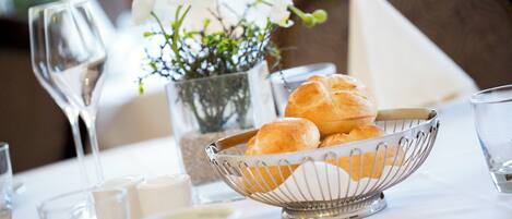 A dining table with a metal bowl containing bread rolls, a vase with white flowers, and several glasses and cups.