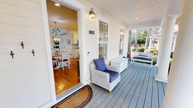 Une véranda avec un fauteuil en osier et un banc, un plancher en bois et une porte donnant sur une cuisine équipée d’une table et de chaises.