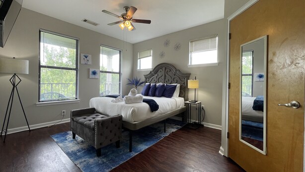 A bedroom with a bed, a ceiling fan, a nightstand, a lamp, a chair, and a large mirror.