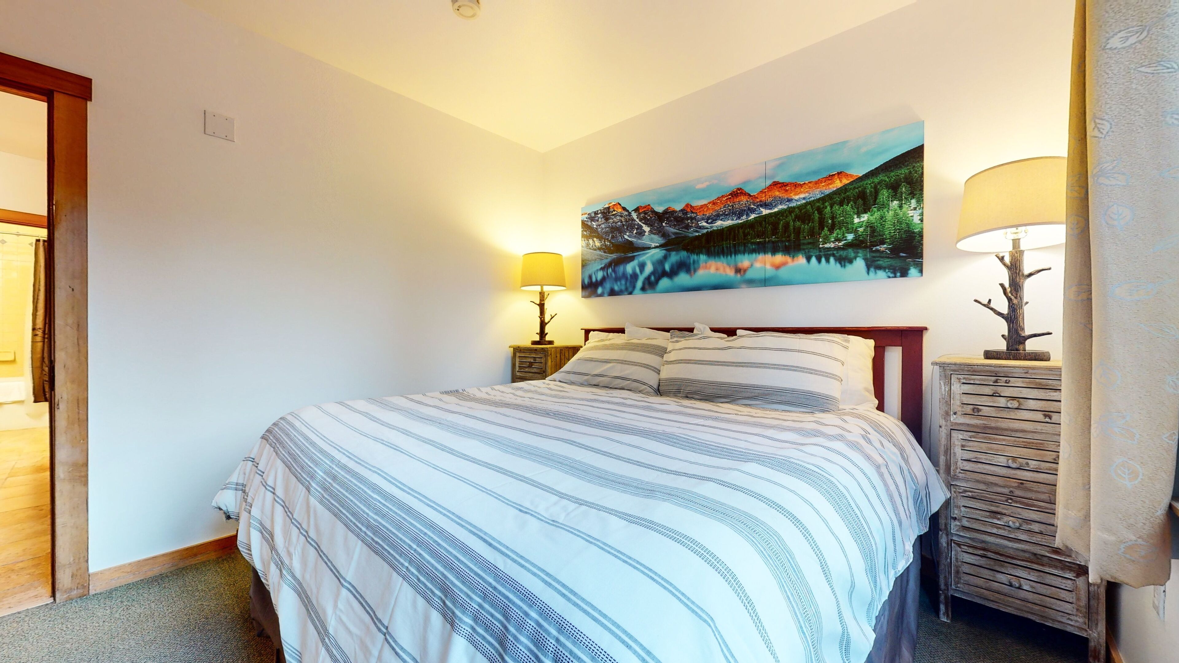 A neatly made bed with striped bedding, a painting of a mountain landscape above it, and two bedside lamps on wooden nightstands.