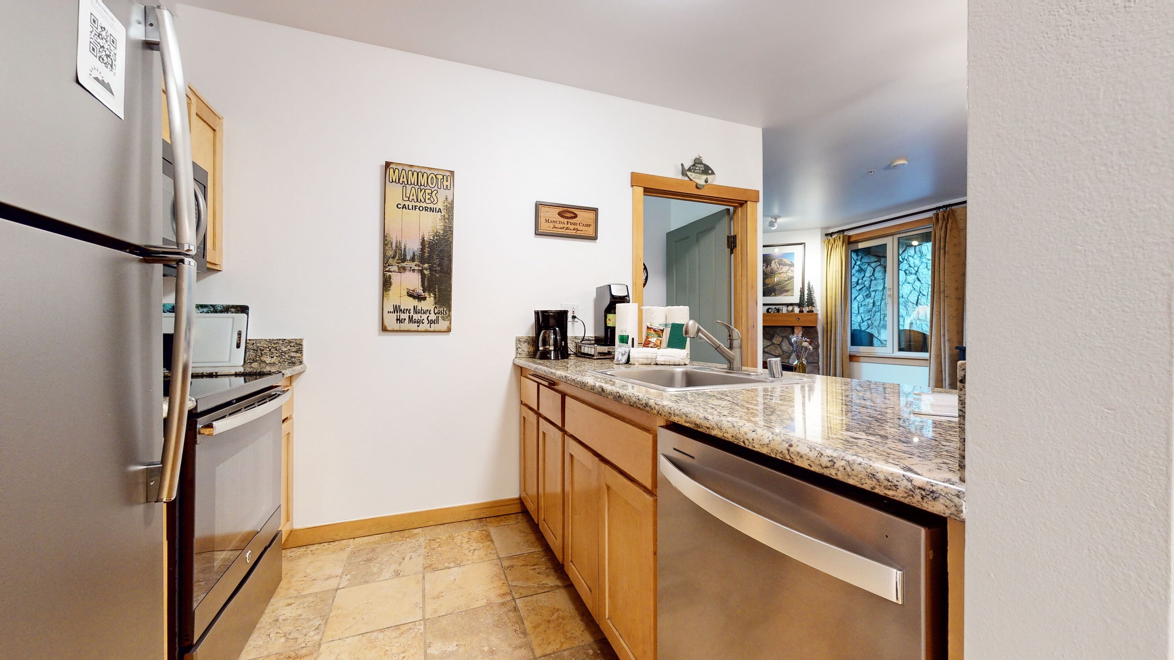A compact kitchen with a refrigerator, stove, and dishwasher. There are two framed pictures on the wall, one of Mammoth Lakes and another of a fish camp. A coffee maker and a bottle of dish soap are on the counter.