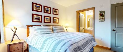 A bedroom with a bed, two bedside tables with lamps, a wooden headboard, and four framed pictures on the wall.