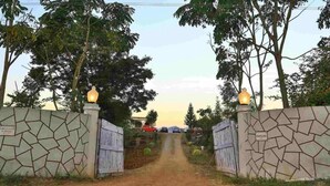 A gated entrance with stone walls and two lit lanterns, leading to a road with parked cars and trees in the background. - Vistara Resort (Araku Valley)