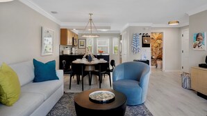 Un salon moderne avec un canapé, un fauteuil, une table basse et un coin repas, la cuisine étant visible au fond.
