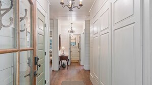 Un couloir aux murs recouverts de panneaux blancs, doté d’un plancher en bois et surmonté d’un lustre. Une porte à panneaux de verre se trouve sur la gauche, et une petite table avec une lampe est placée au centre.