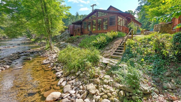 A wooden cabin with a glass front, surrounded by lush greenery and a stream. - Evans Cabin Creekside Cabin Rental (Bryson City)