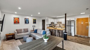 A modern living room with a sofa, coffee table, and a dining table. There is a kitchen area with cabinets and appliances.