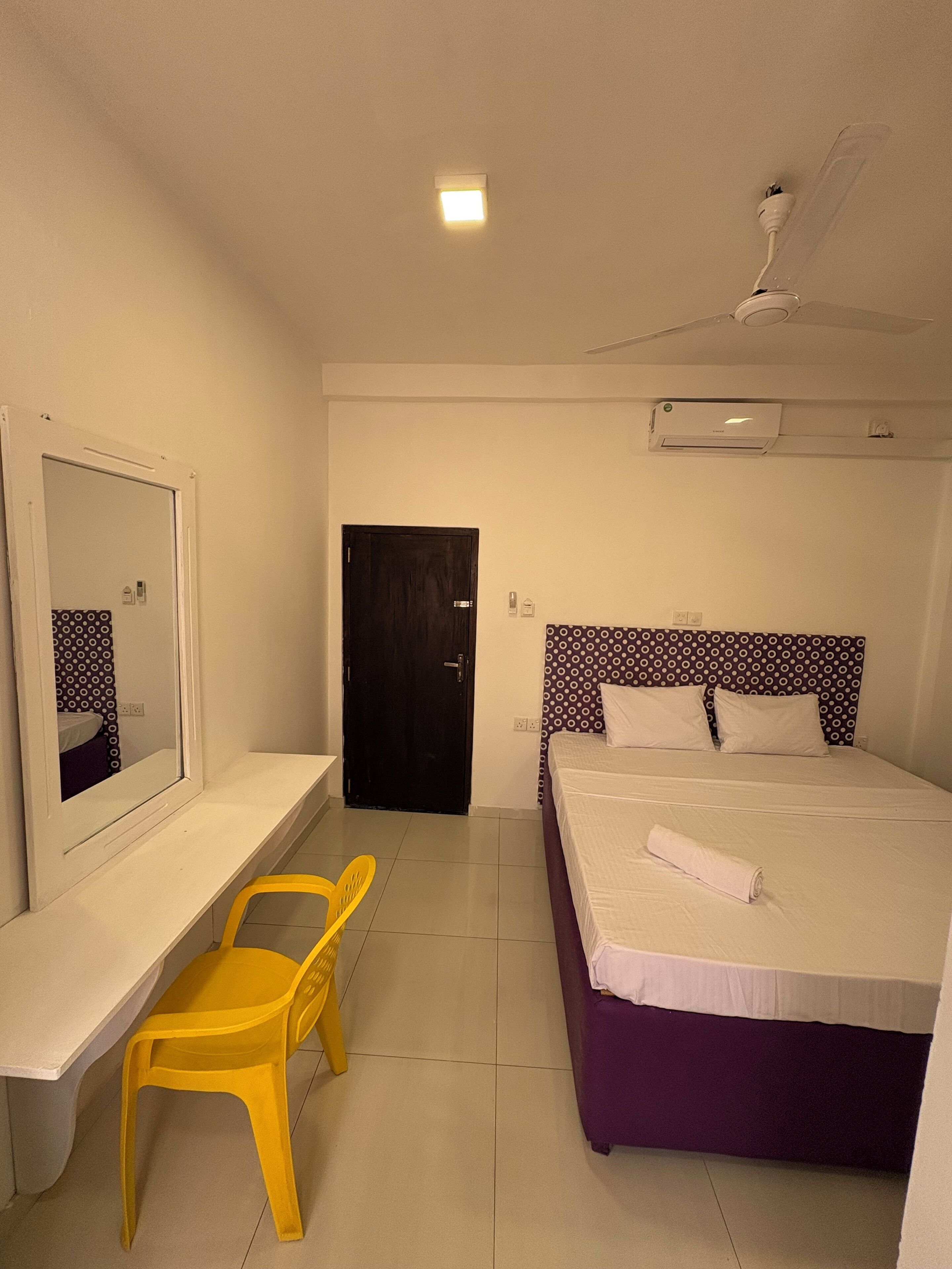 A hotel room with a bed, a desk, a chair, a mirror, a door, and an air conditioning unit.