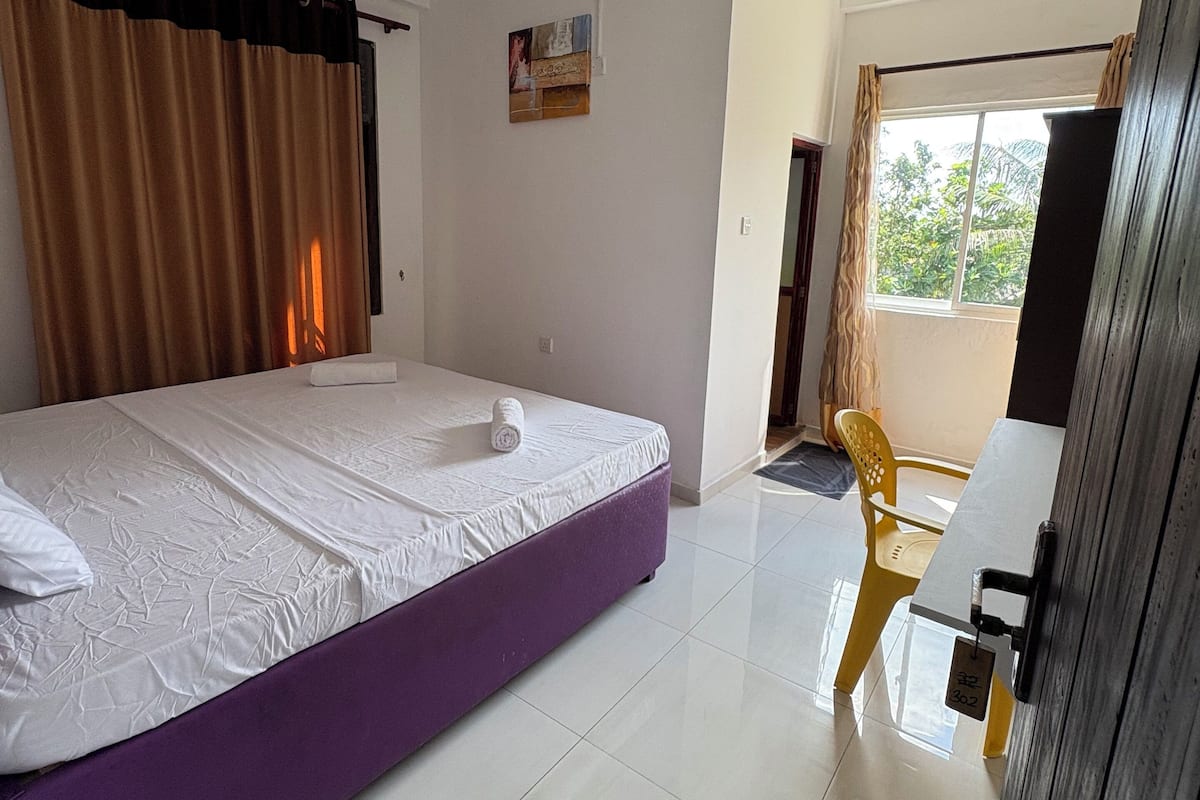 A hotel room with a bed, a ceiling fan, a window with curtains, and a door leading to another room.