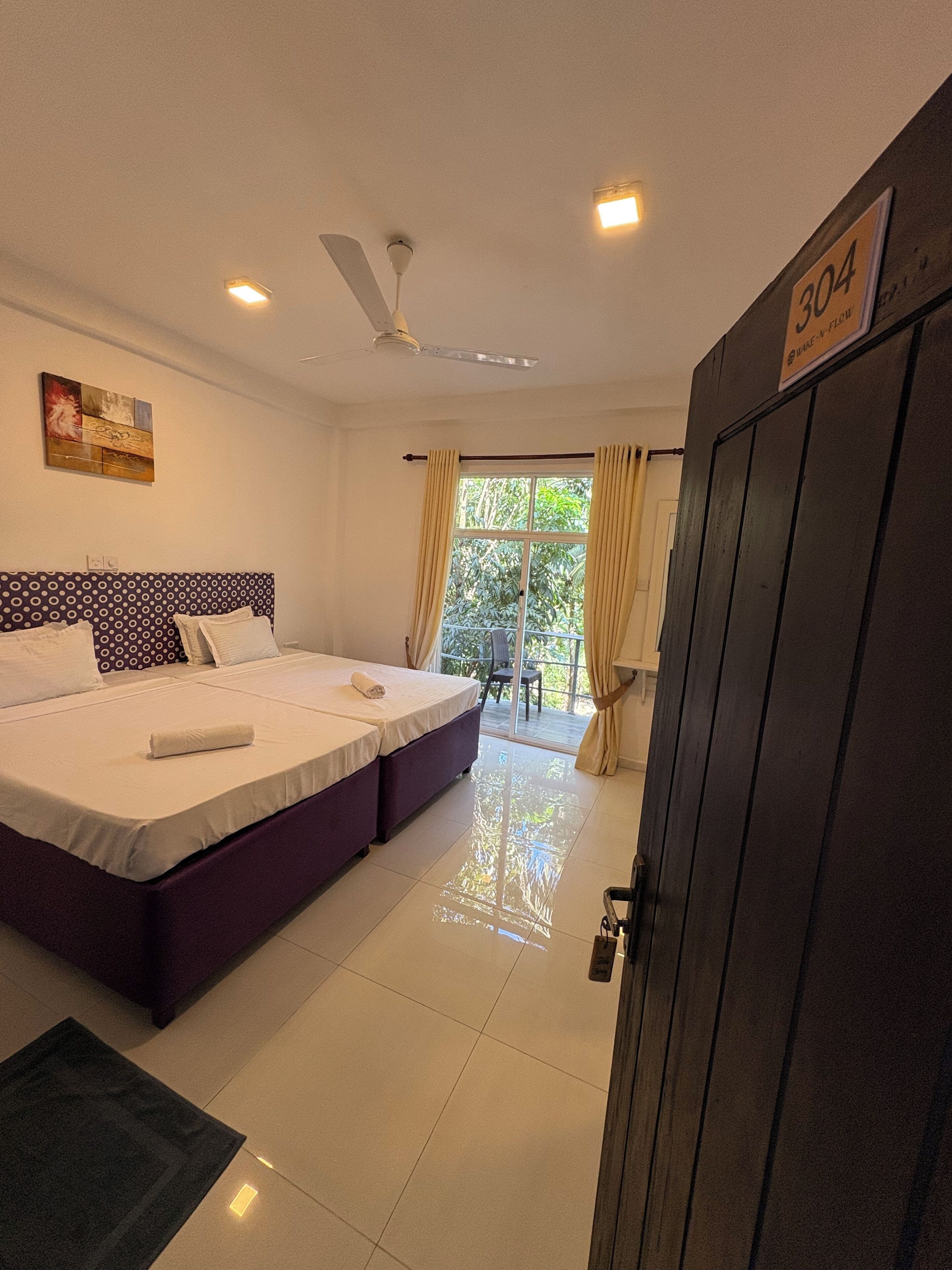 A hotel room with a bed, a ceiling fan, a window with curtains, and a door with a number sign.