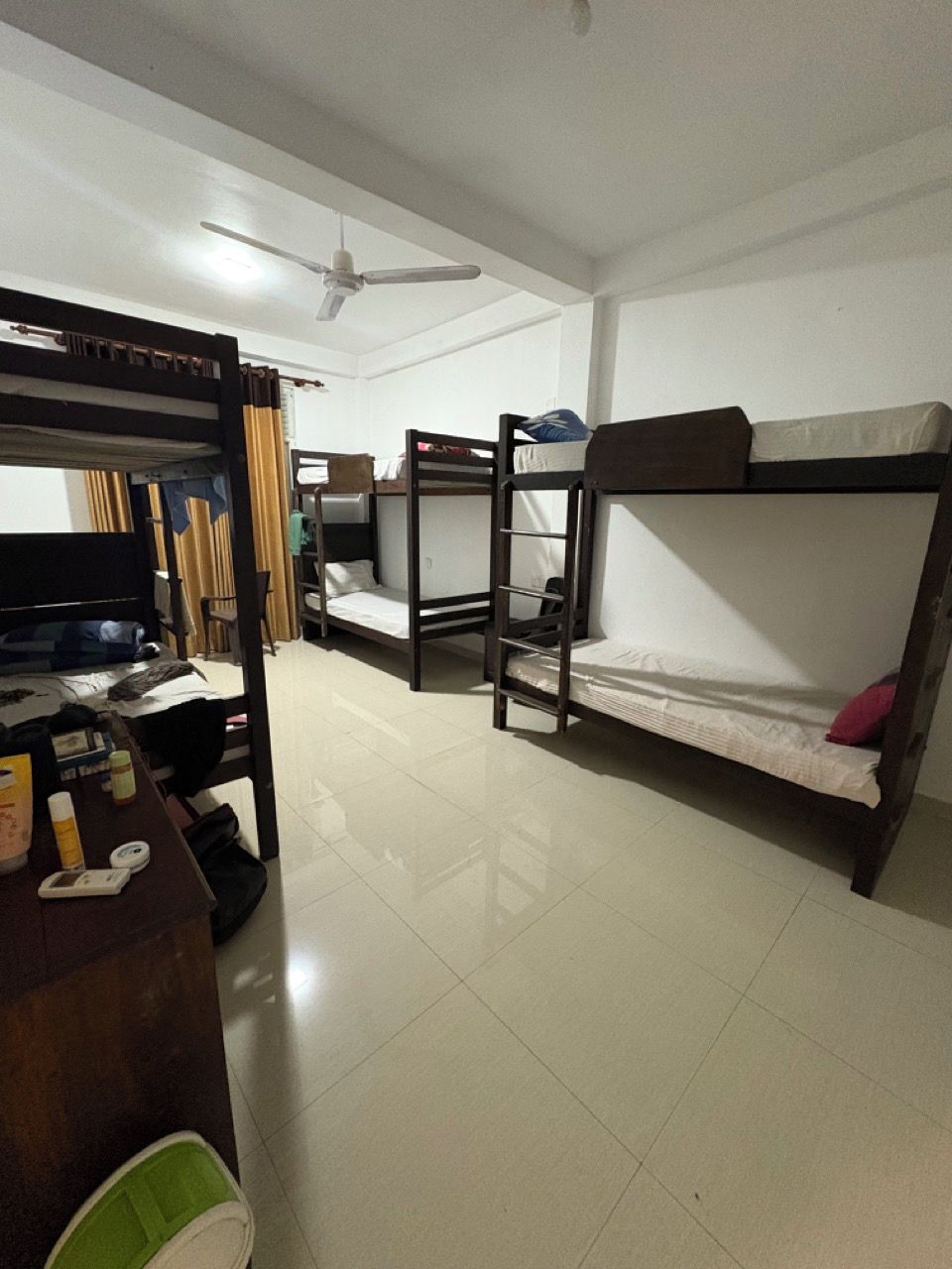 A dormitory room with bunk beds, a desk with personal items, and a ceiling fan.