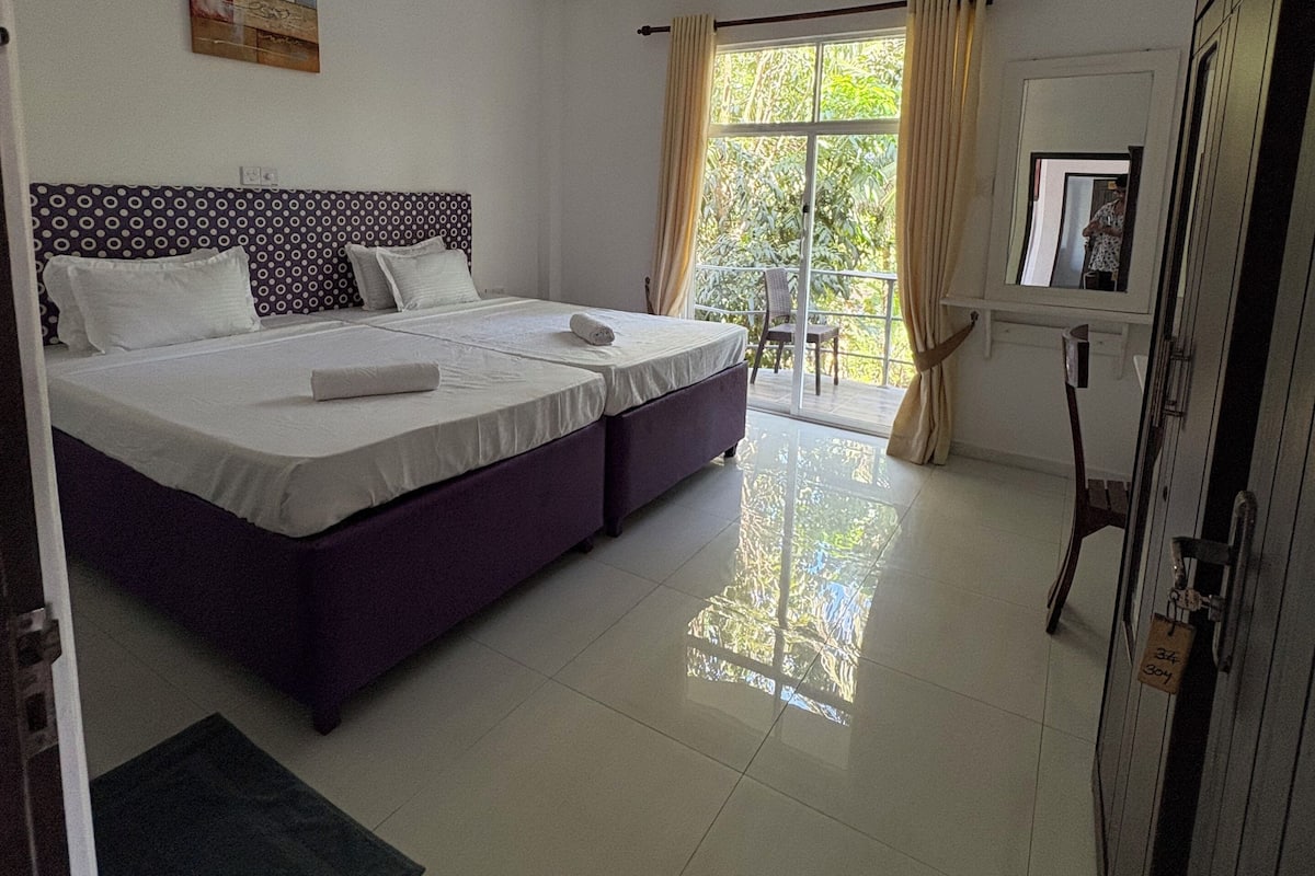 A bedroom with two beds, a ceiling fan, a window with curtains, and a view of greenery outside.
