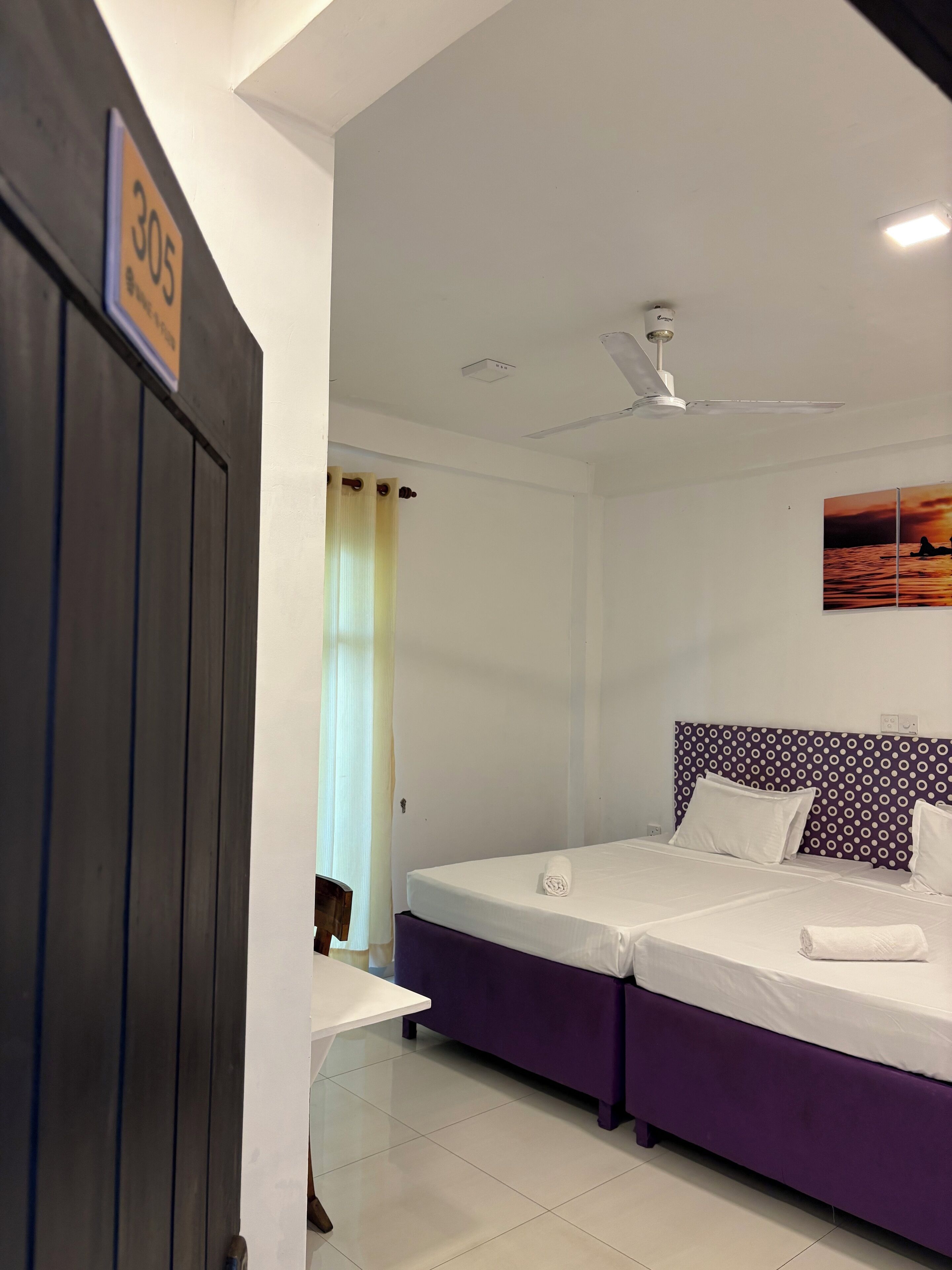 Une chambre d’hôtel avec deux lits, un ventilateur de plafond, un tableau représentant un coucher de soleil et une pancarte indiquant le numéro de chambre : 305.
