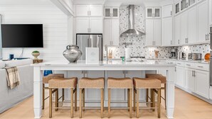 Eine moderne Küche mit weißen Schränken, einem Edelstahlkühlschrank, einer großen Kochinsel mit Barhocker und einem flachbildfernseher an der Wand.