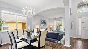 Un salon moderne avec une table à manger en verre, des sièges confortables, un lustre et une vue sur l’extérieur.