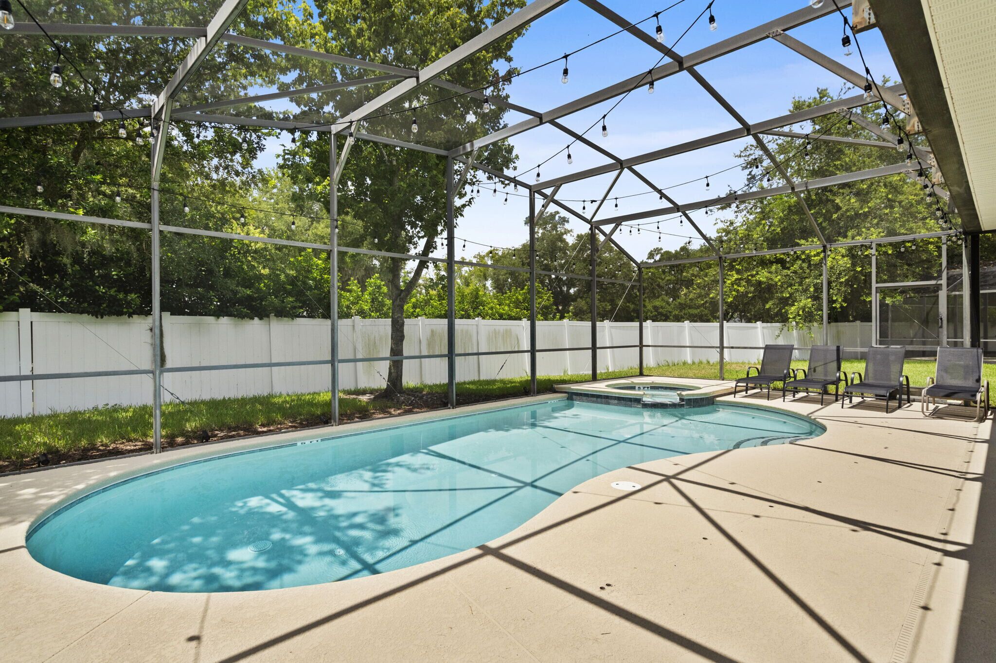 A covered outdoor swimming pool with a white fence, lounge chairs, and string lights.