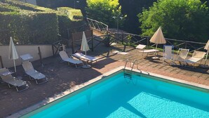 Une piscine entourée d’une terrasse en briques et de verdure, avec des chaises longues et des parasols.