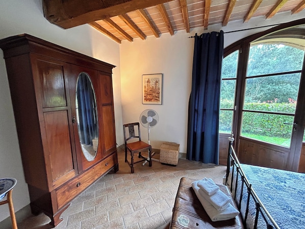 Um quarto com um armário de madeira, uma cadeira, um ventilador, uma janela com cortinas e um banco com toalhas.