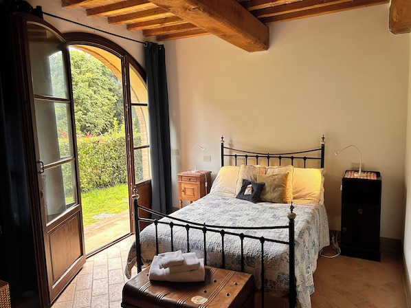 A bedroom with a bed, a bedside table, a lamp, and a view of greenery through a window.