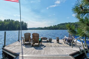 A wooden dock with chairs and a table, overlooking a calm lake with a forested shoreline.