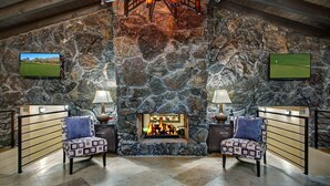 Habitación de paredes de piedra con chimenea, dos sillones y una pantalla de televisión mostrando un campo de golf.