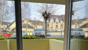 Une vue depuis une fenêtre donnant sur un espace recouvert d’un gazon synthétique vert, un mur jaune et le reflet d’un immeuble doté de nombreuses fenêtres et de balcons.