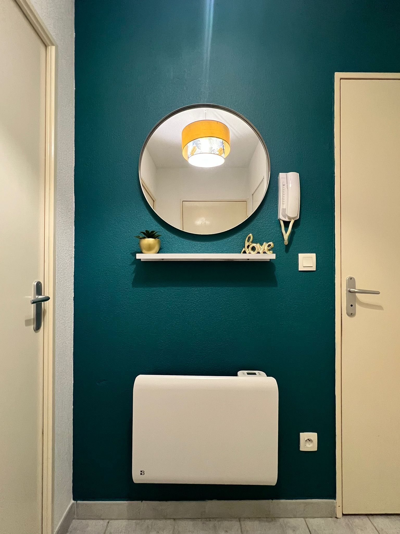 Een badkamer met een blauwgroene muur, een ronde spiegel met geel licht, een witte telefoon, een klein plankje met een plant en een decoratie met de tekst 'love', en een witte radiator.